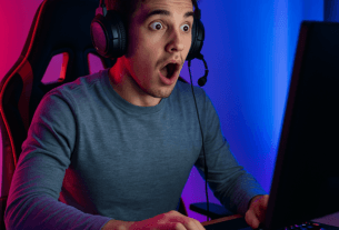 Um gamer surpreso sentado em sua cadeira gamer, usando fones de ouvido e jogando no computador. A cena é iluminada por luzes coloridas, criando uma atmosfera imersiva.